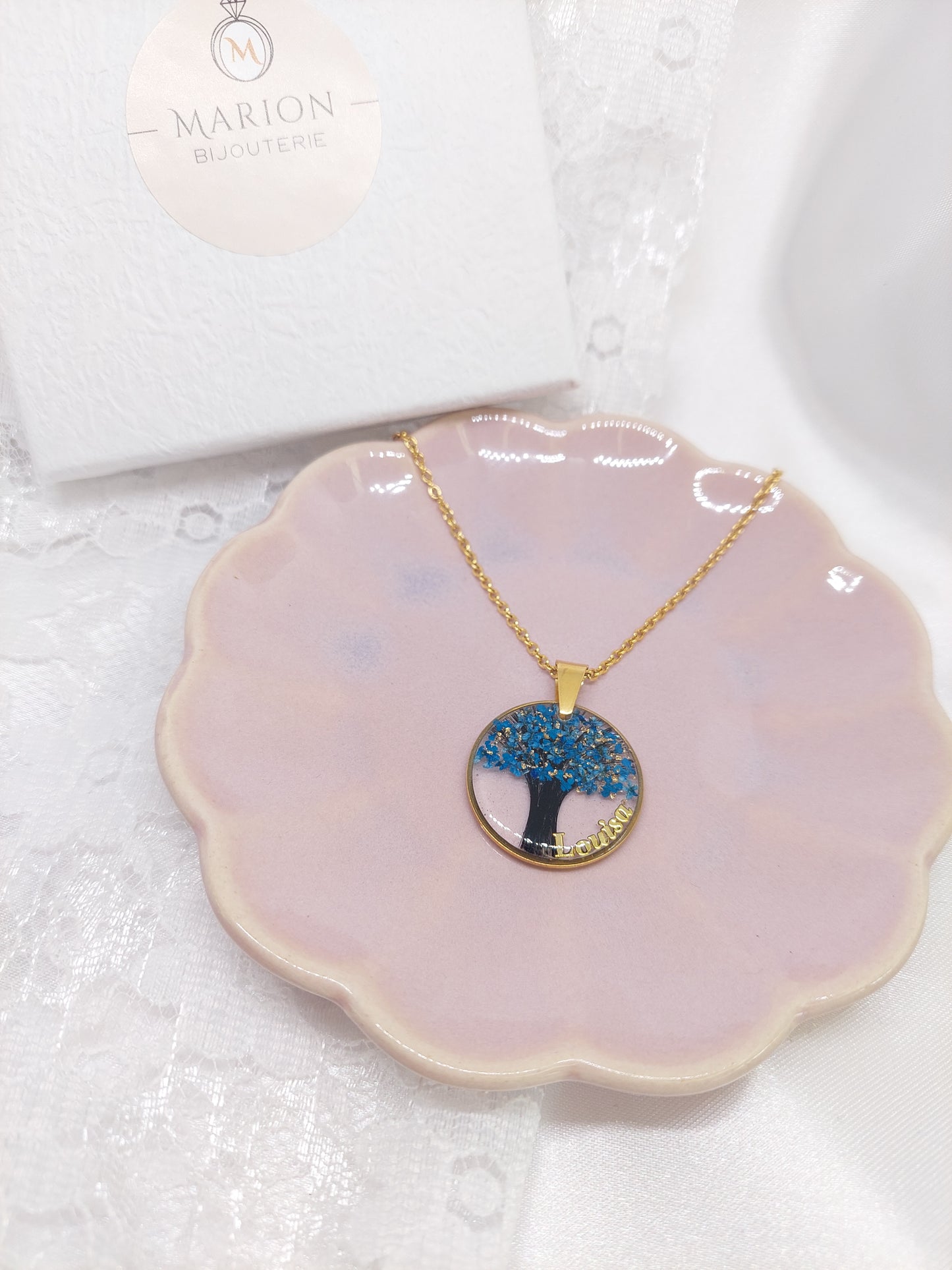 bijou souvenir: collier avec un pendentif rond et une mèche de cheveux mise à l'intérieur en forme d'arbre de vie avec des fleurs bleus et des feuilles d'argent ainsi qu'un prénom sur le côté