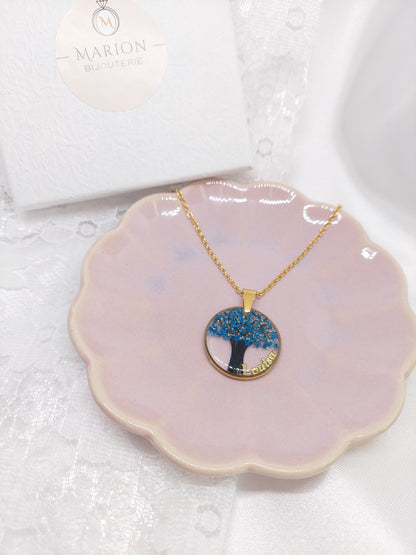 bijou souvenir: collier avec un pendentif rond et une mèche de cheveux mise à l'intérieur en forme d'arbre de vie avec des fleurs bleus et des feuilles d'argent ainsi qu'un prénom sur le côté